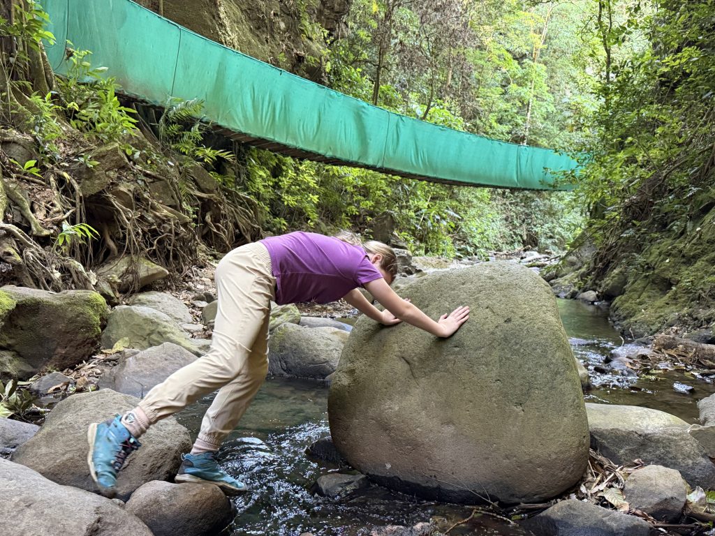 A child leans forward with both hands on a large boulder in a shallow forest stream, bracing one foot on a rock as if pushing it. A green-covered suspension bridge spans the rocky ravine above, surrounded by dense vegetation.
